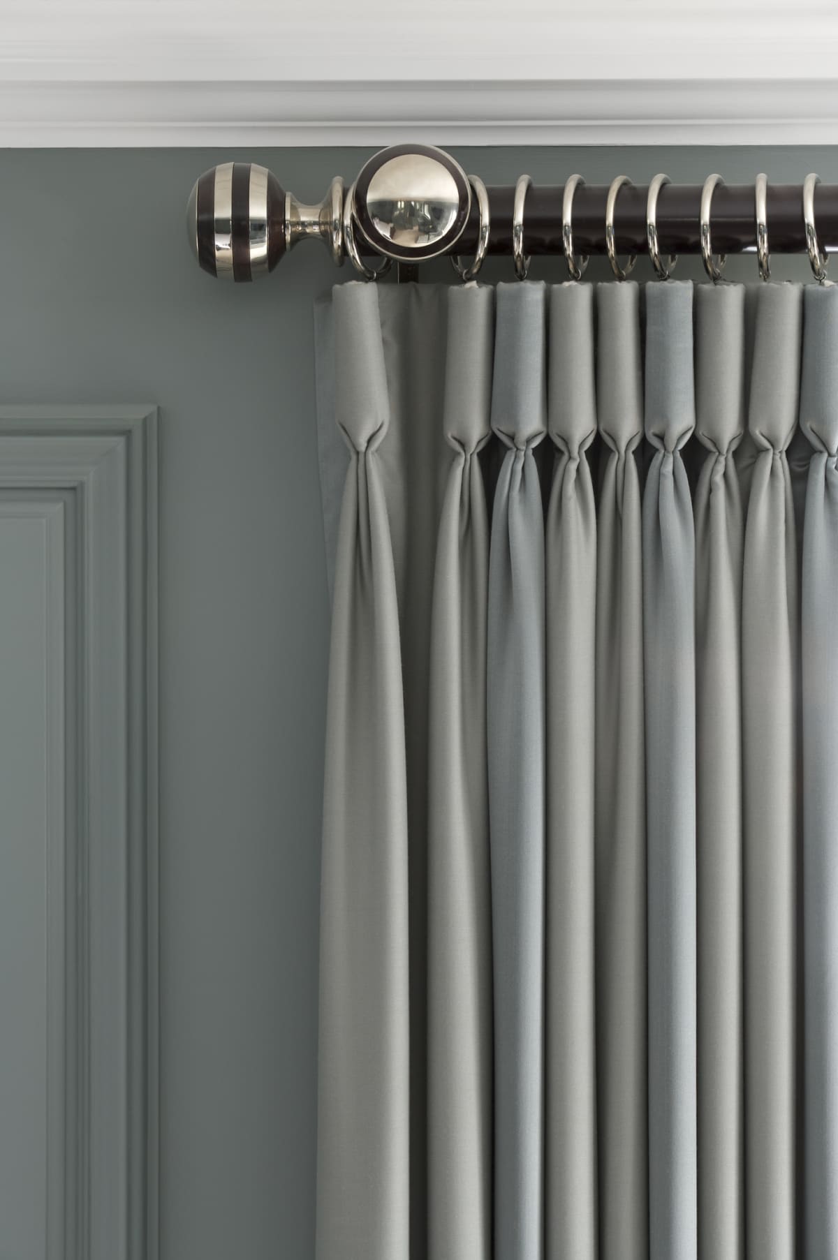 Grey pinch pleated curtains hanging from a wood and metal curtain rail with metal rings.