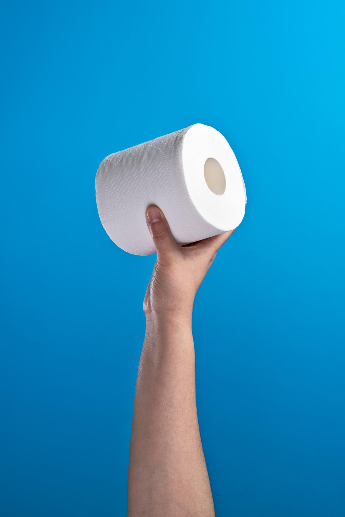 A hand holding a single roll of toilet paper against a blue background