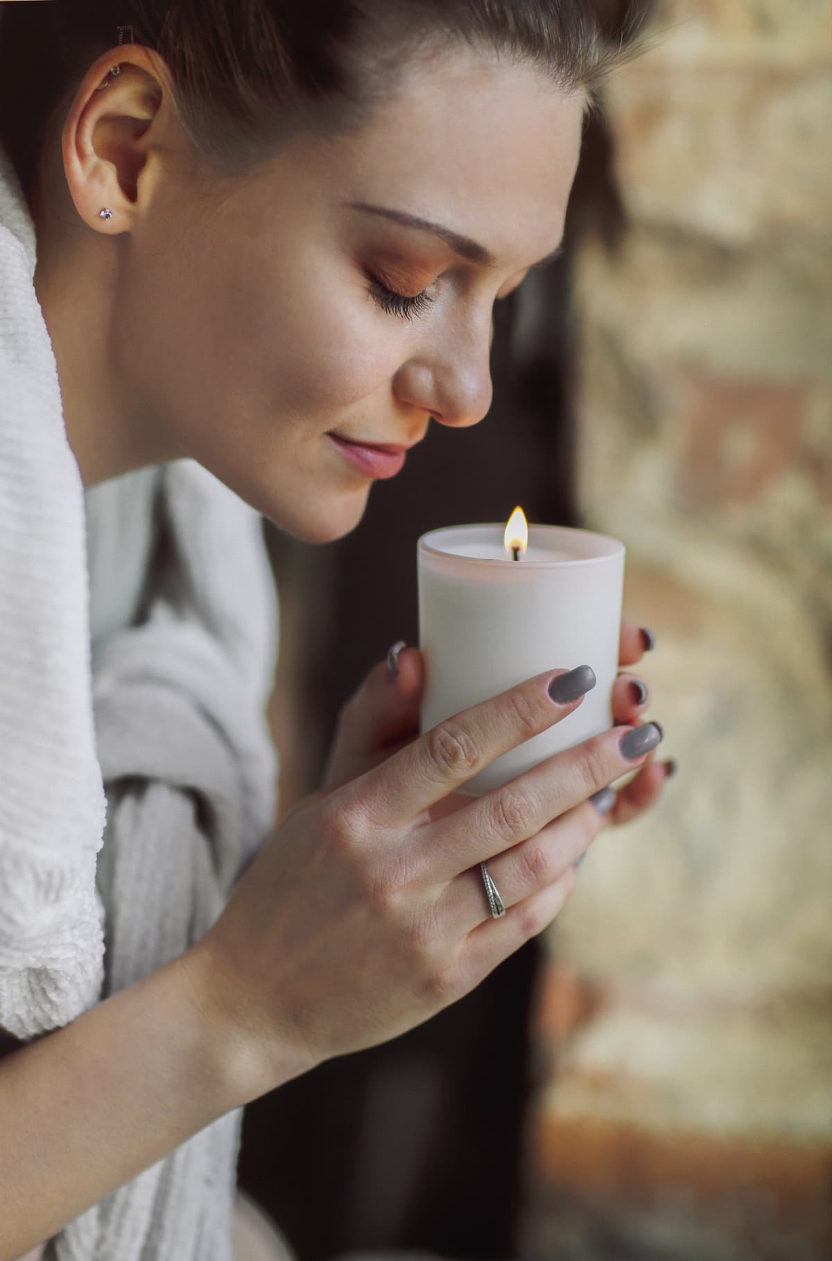 Side portrait of young calm woman holding lit candle in hands, closing eyes bringing close to her face and inhaling its aroma, smelling familiar scent while enjoying happy moment of comfort
