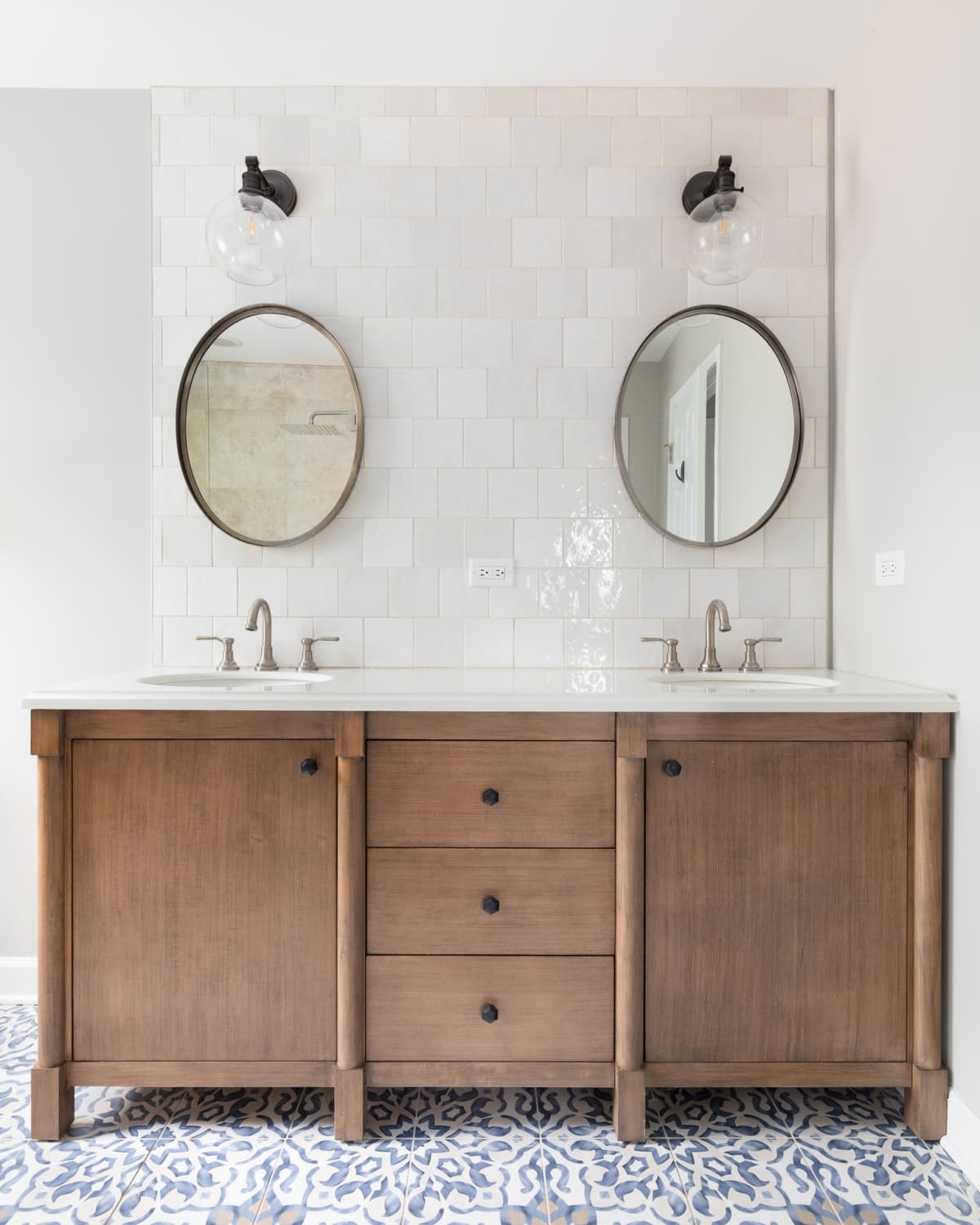 A cozy bathroom with a patterned tile floor, natural wood vanity, tiled backsplash, and lights mounted above circular mirrors.