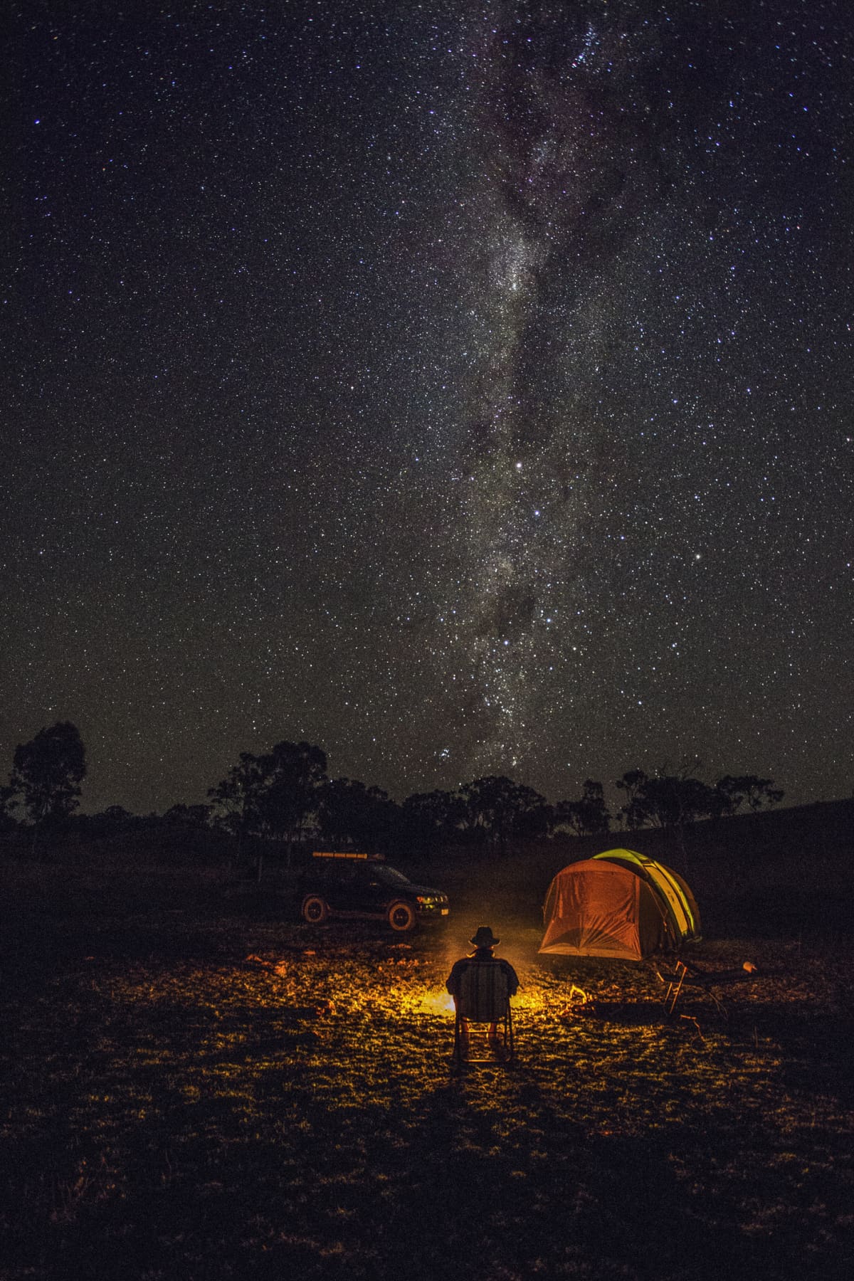 man sits in a folding chair next to a campsite under a sky filled with stars