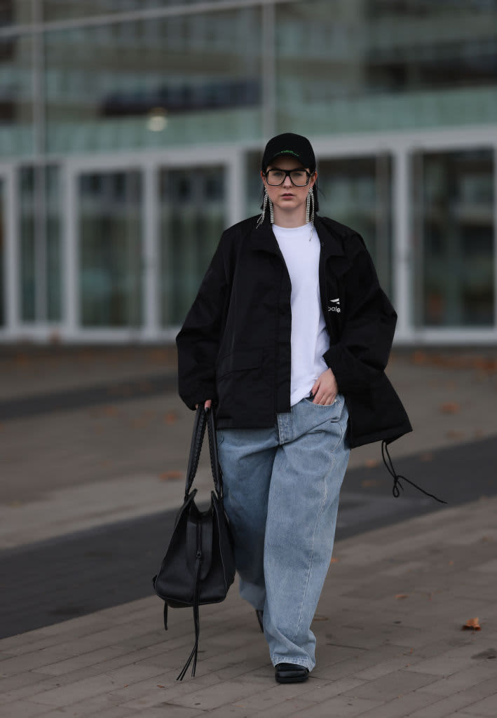Woman walking in baggy jeans, a white t-shirt, and black jacket carrying a purse and wearing a hat and black-rimmed glasses