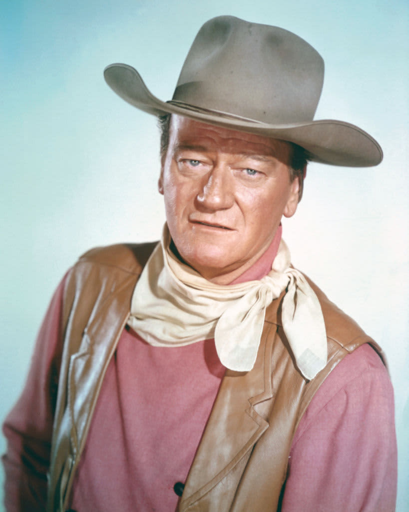 John Wayne (1907 - 1979), US actor wearing a tan leather waistcoat, a pink shirt and a white neckerchief, in a studio portrait, against a light blue background, circa 1970. (Photo by Silver Screen Collection/Getty Images)