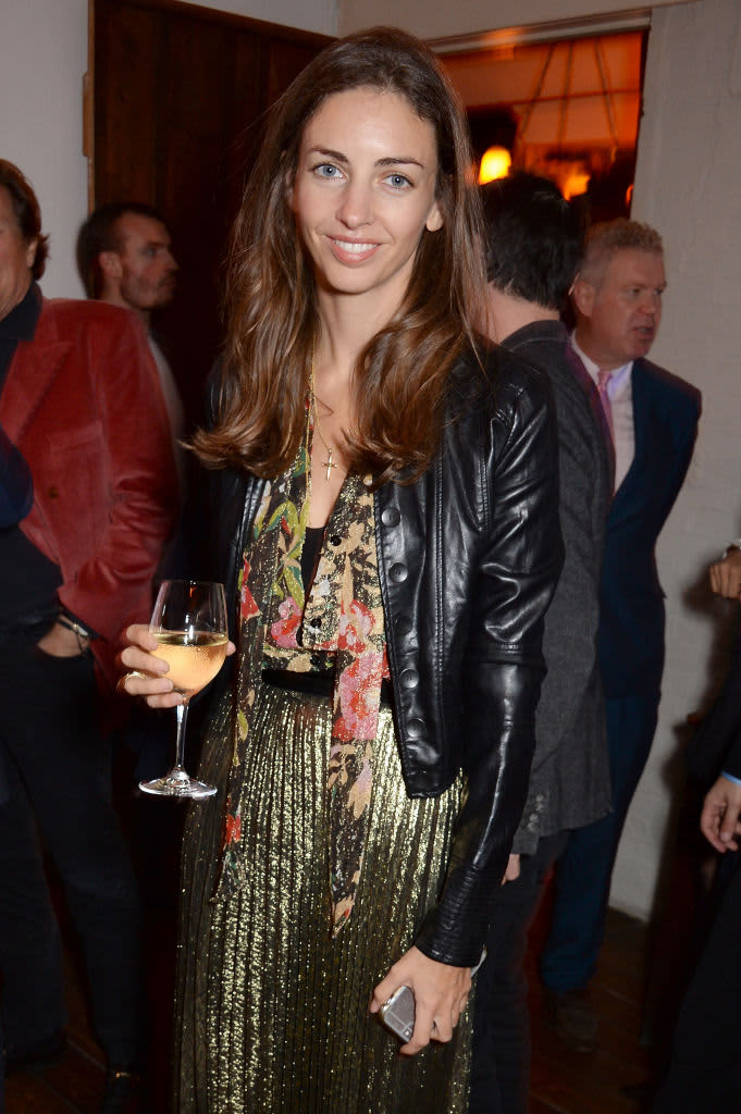 A smiling Rose Hanbury attends an event with gold lame pants, a black shirt and a colorful scarf with a black leather jacket, holding a glass of wine and her phone