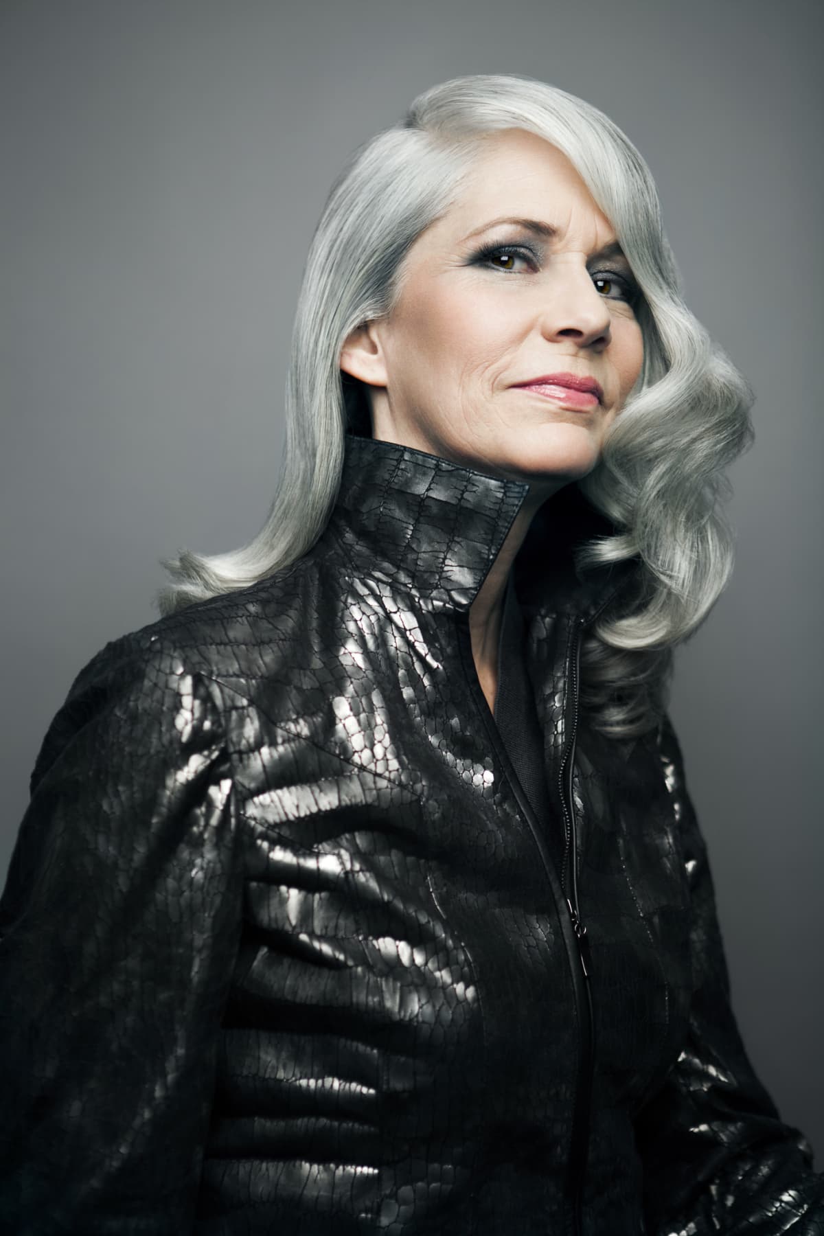 Mature woman with long, silvery, grey hair wearing a black leather jacket in front of a grey background