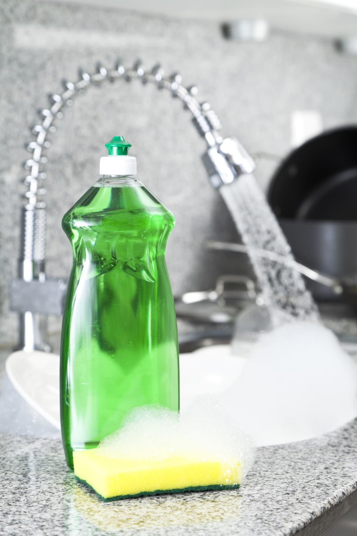 Green Dishwashing Detergent Bottle and Sponge with Kitchen Sink Full of Dirty Dishes in Background