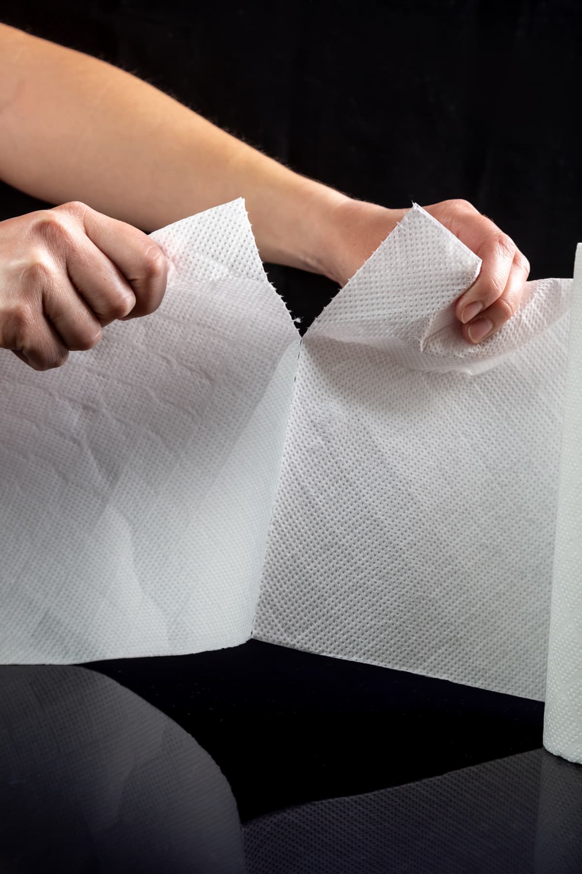 Hands tear off a piece of white paper towel from a roll on a black background. Vertical orientation. High quality photo.