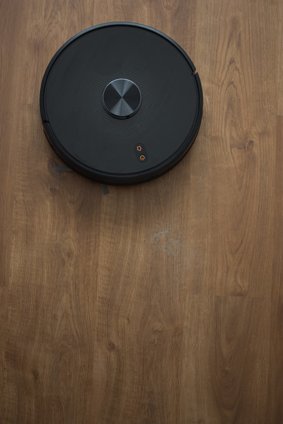 black robot vacuum cleaner cleaning dust and sand on a wooden laminate floor, displaced at the top with orange power LEDs · vertical view