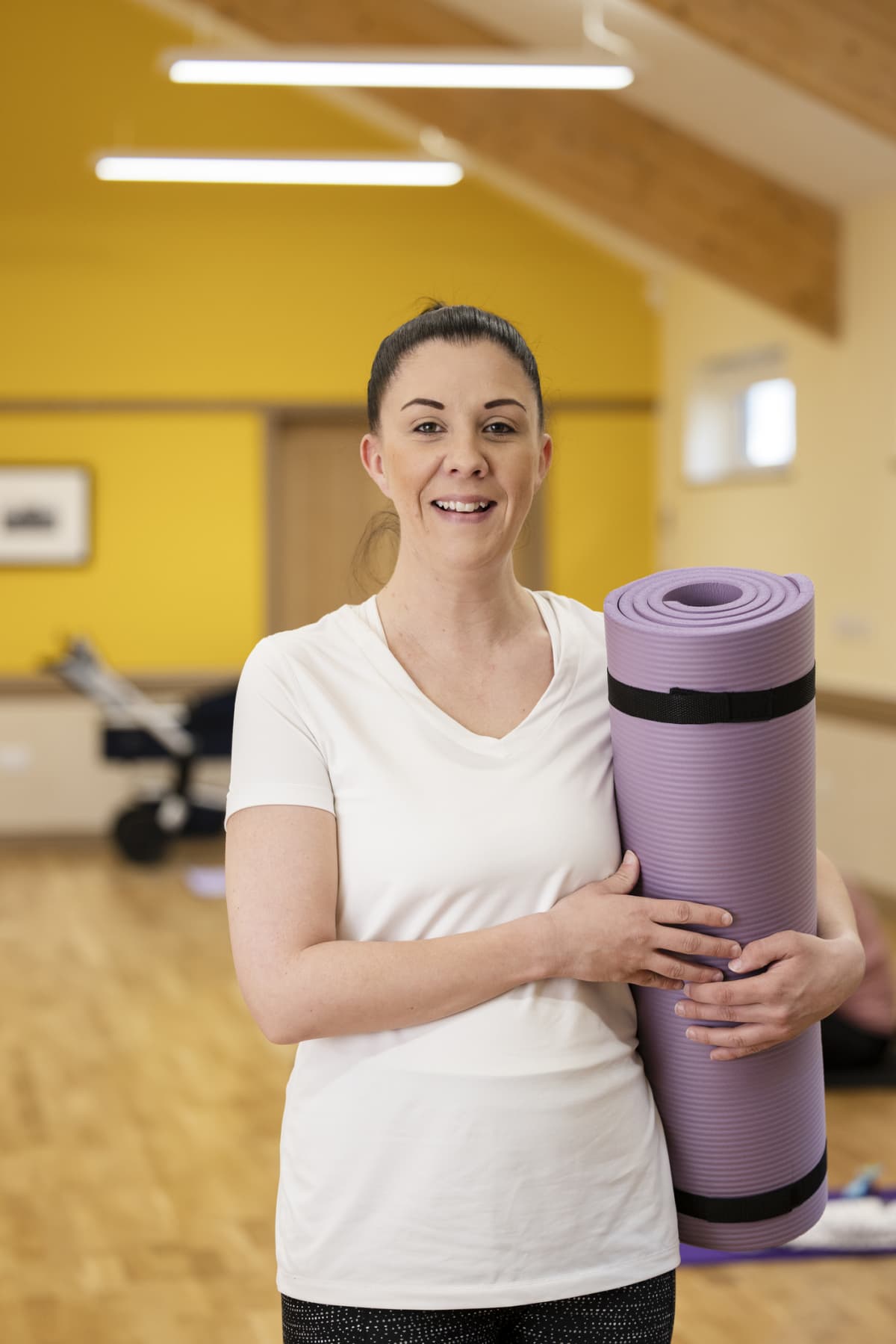 A front-view waist-up portrait shot of a woman in a yoga class, she is standing, smiling, and looking at the camera while holding an exercise mat.